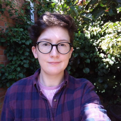 Jake is a white transmasculine person with short brown hair and round dark-rimmed glasses. They are smiling at the camera, in front of greenery.